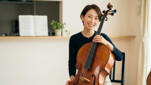 チェロを愛おしそうに抱える日本人女性と、明るく片付けられた自宅練習スペース。